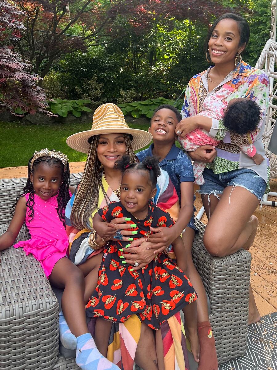 Simone I. Smith sits outdoors in a wide straw hat with four of her grandchildren gathered around her, one daughter kneeling alongside holding a baby.