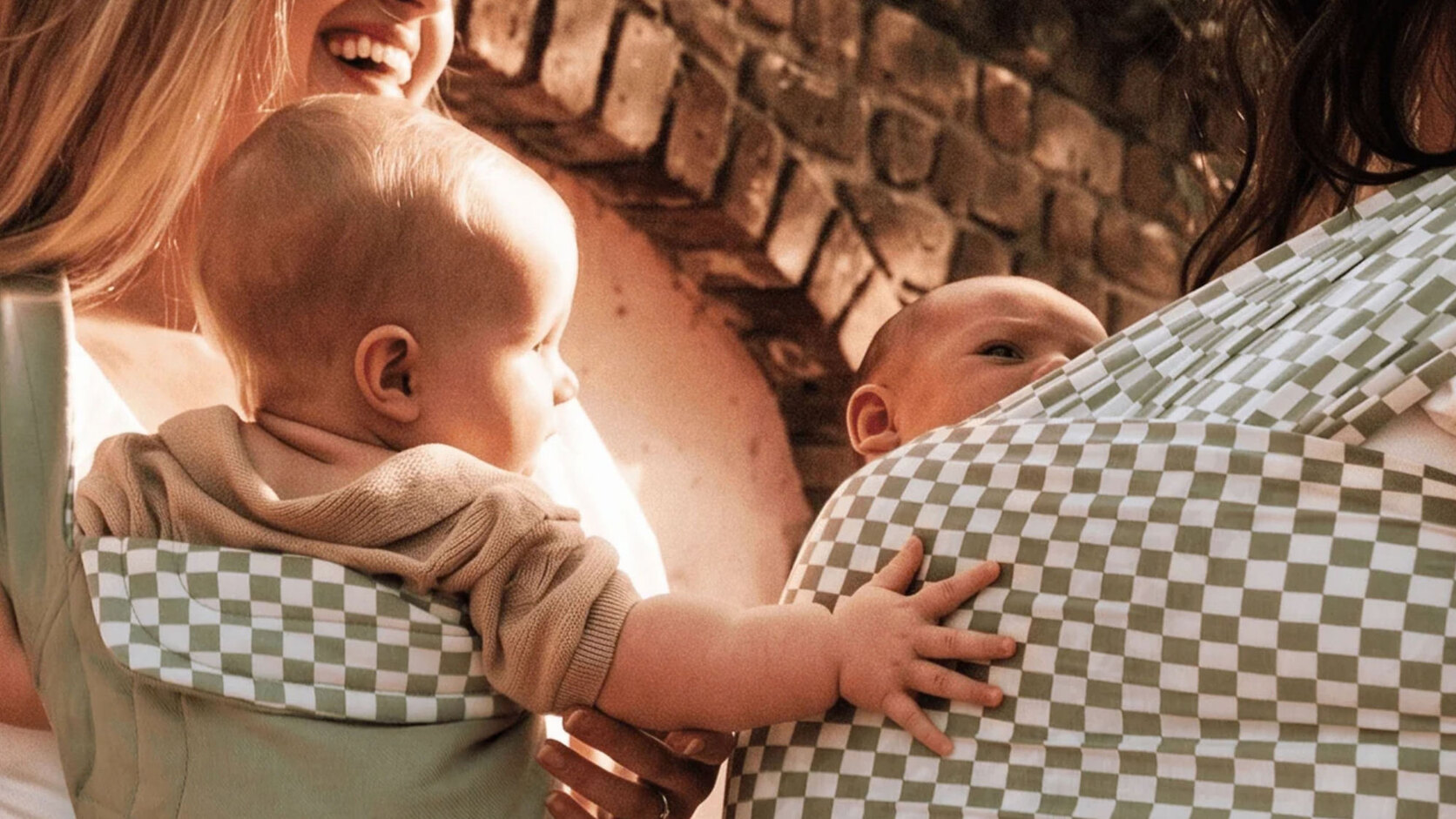 Two mothers carry their babies in green-and-white checkered Solly wraps against a sunlit brick wall, the babies' hands reaching toward one another.