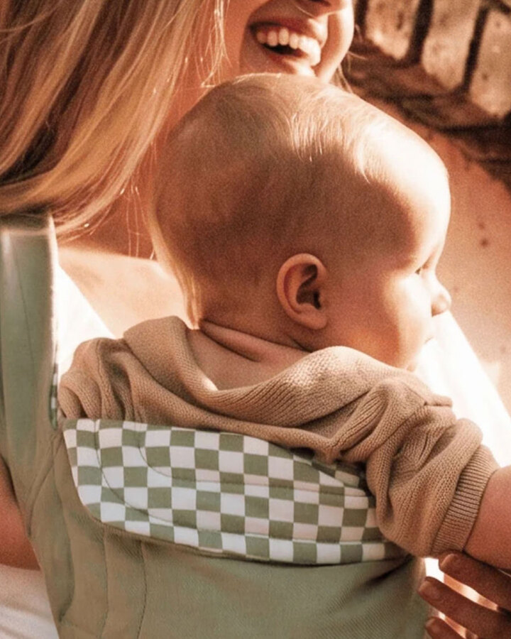 Two mothers carry their babies in green-and-white checkered Solly wraps against a sunlit brick wall, the babies' hands reaching toward one another.
