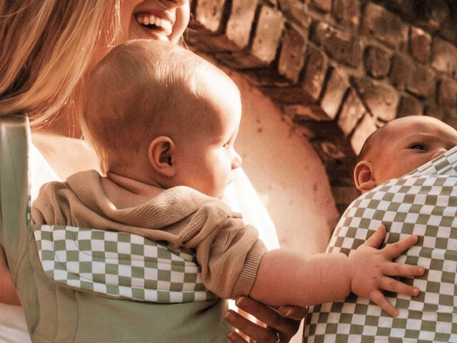 Two mothers carry their babies in green-and-white checkered Solly wraps against a sunlit brick wall, the babies' hands reaching toward one another.
