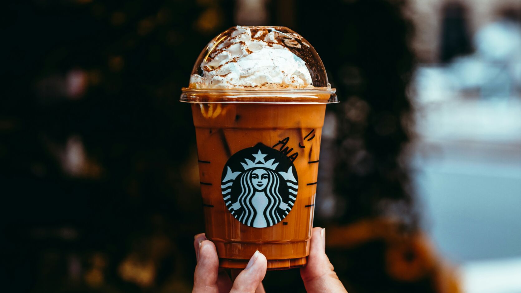 A hand holds up an iced Starbucks pumpkin drink, whipped cream dusted with spice under a clear dome lid, against a softly blurred autumn street.
