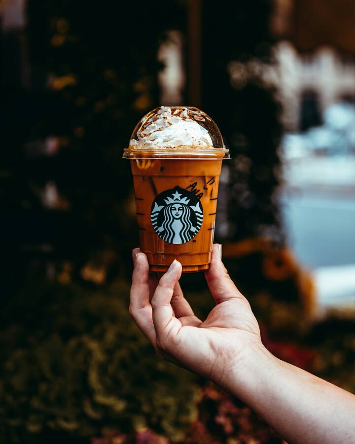 A hand holds up an iced Starbucks pumpkin drink, whipped cream dusted with spice under a clear dome lid, against a softly blurred autumn street.