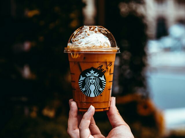 A hand holds up an iced Starbucks pumpkin drink, whipped cream dusted with spice under a clear dome lid, against a softly blurred autumn street.