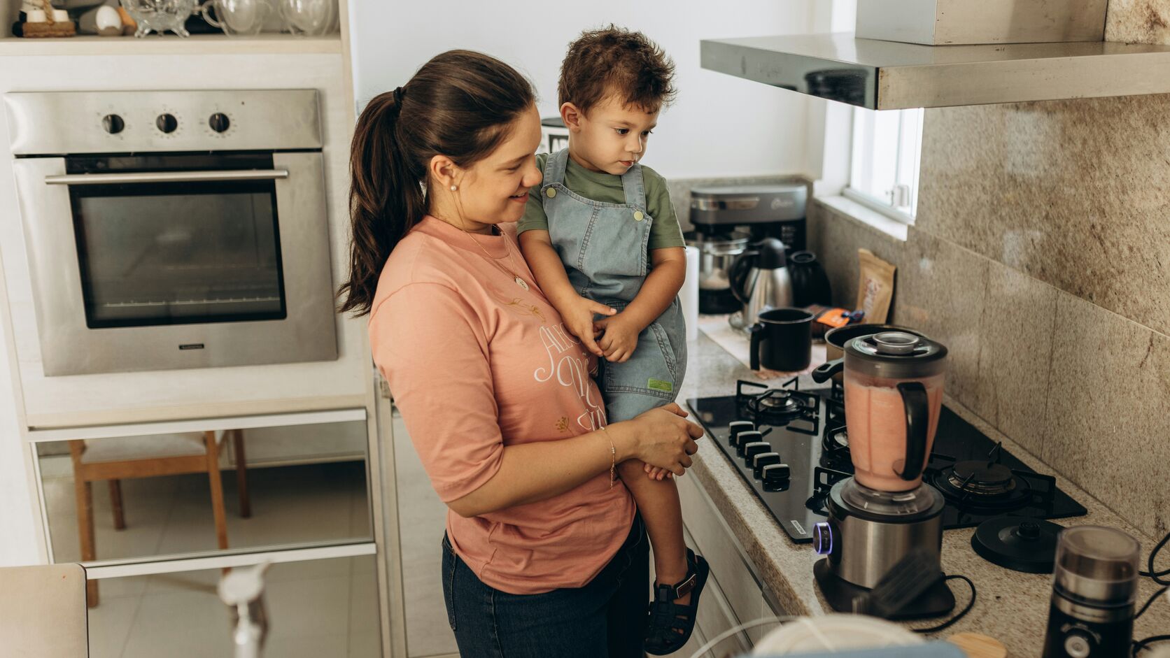A smiling mother holds her toddler on her hip in a bright kitchen, a blender of pink puree on the counter beside the stove.