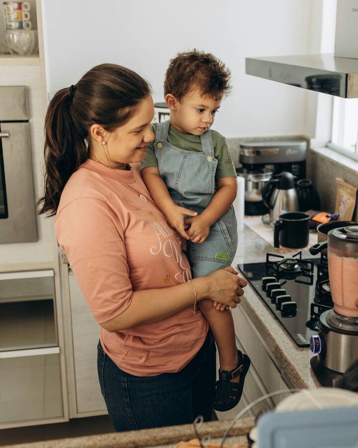 A smiling mother holds her toddler on her hip in a bright kitchen, a blender of pink puree on the counter beside the stove.