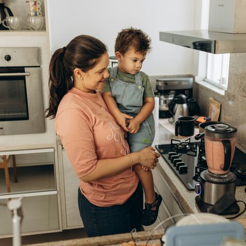 A smiling mother holds her toddler on her hip in a bright kitchen with a blender of pink puree on the counter.
