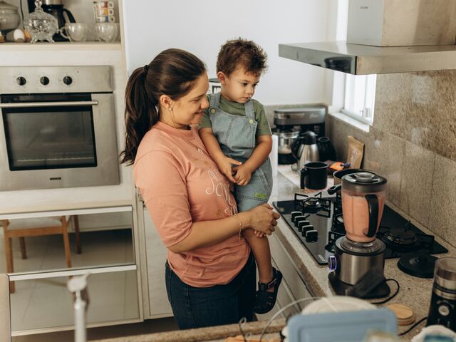 A smiling mother holds her toddler on her hip in a bright kitchen with a blender of pink puree on the counter.