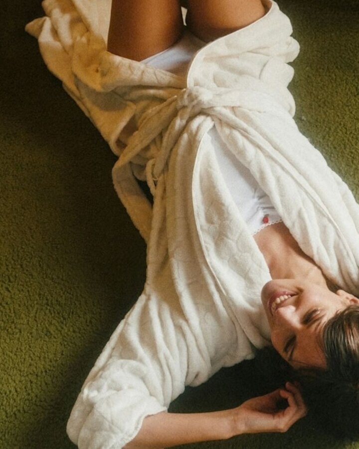 A woman in a cream robe lies relaxed on a green carpet, smiling, one arm tucked behind her head, in warm natural light.