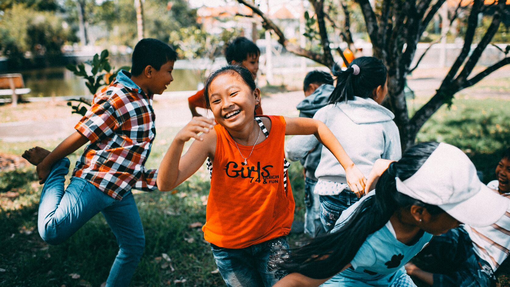 A group of children run and play together on the grass in a park, a laughing girl in an orange tank top at the center of the game.