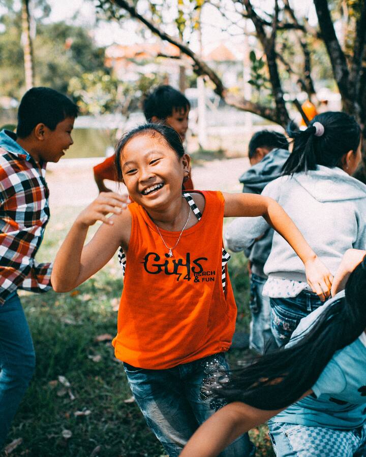 A group of children run and play together on the grass in a park, a laughing girl in an orange tank top at the center of the game.