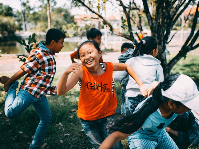 A group of children run and play together on the grass in a park, a laughing girl in an orange tank top at the center of the game.