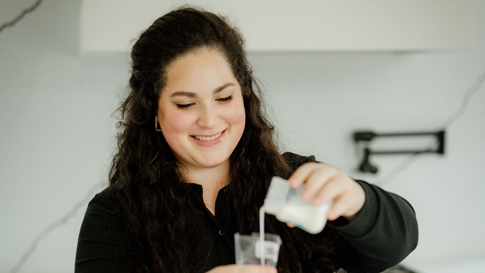 Tamari Jacob smiles as she pours freshly pumped breast milk into a storage bag at a white marble kitchen counter.