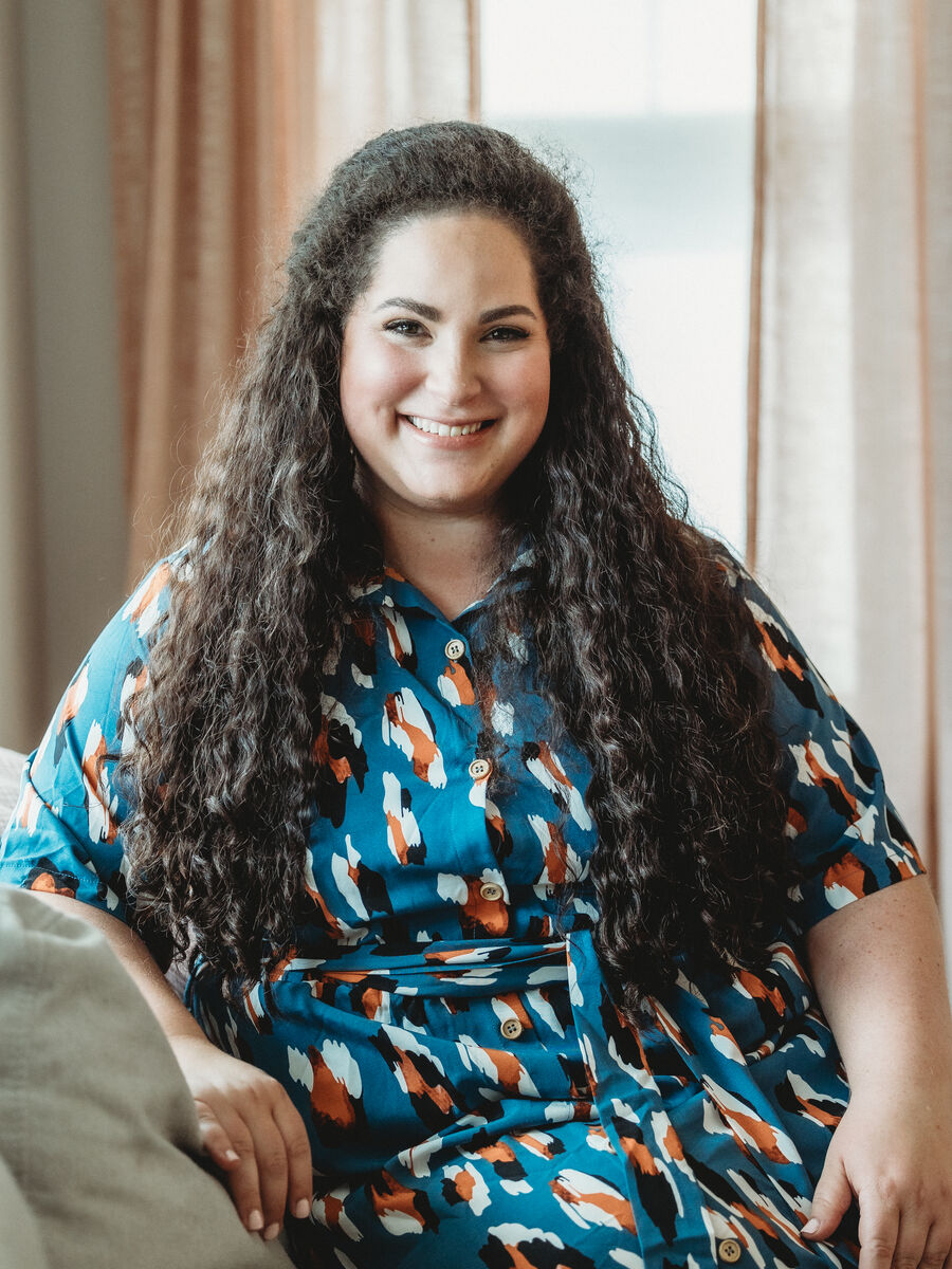 Tamari Jacob sits on a sofa in a teal patterned shirt dress, smiling in front of soft blush curtains.