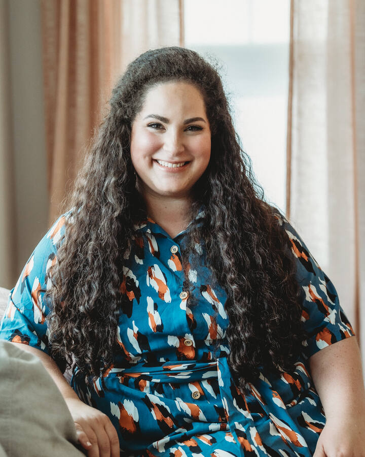 Tamari Jacob sits on a sofa in a teal patterned shirt dress, smiling in front of soft blush curtains.