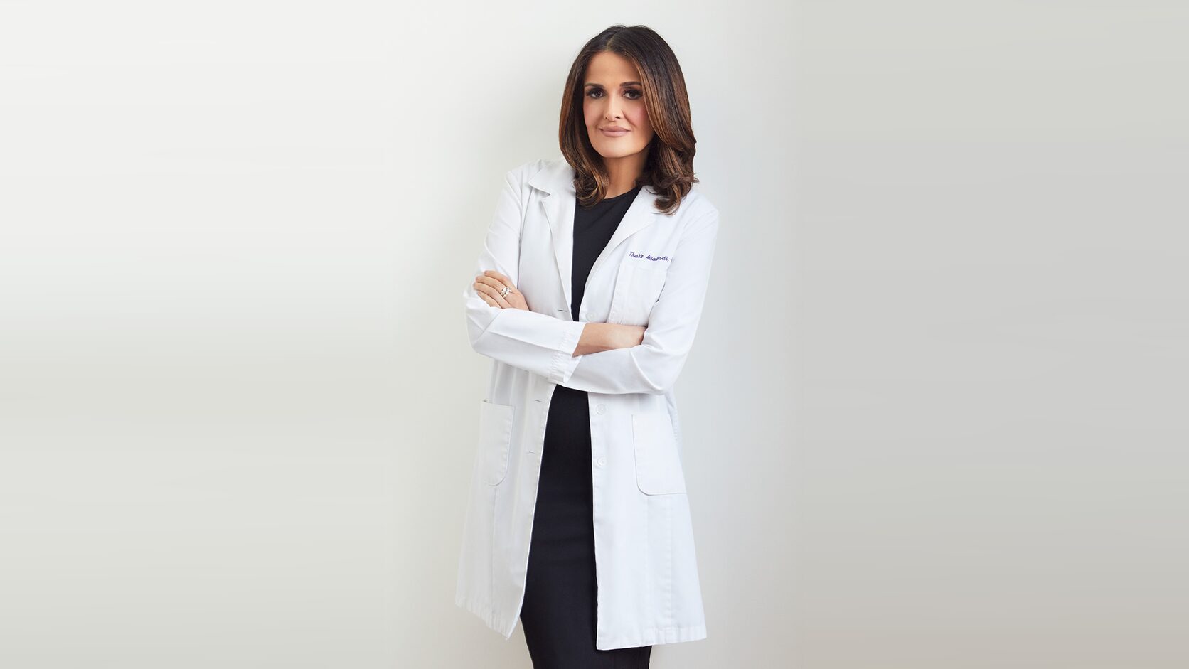 Dr. Thaïs Aliabadi stands against a pale grey studio backdrop in a white lab coat over a black dress, arms folded, looking at the camera.