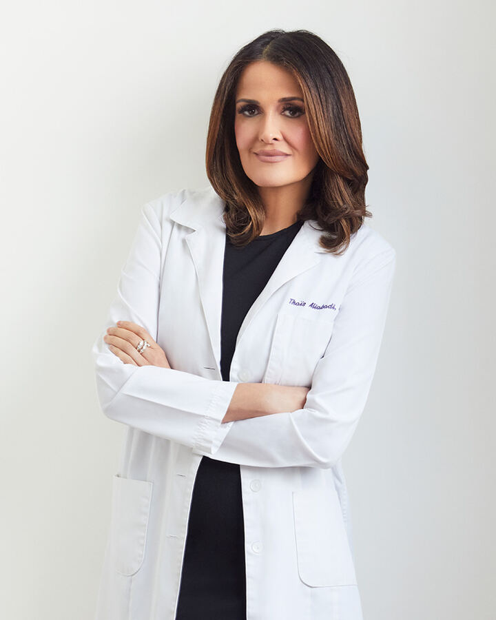 Dr. Thaïs Aliabadi stands against a pale grey studio backdrop in a white lab coat over a black dress, arms folded, looking at the camera.