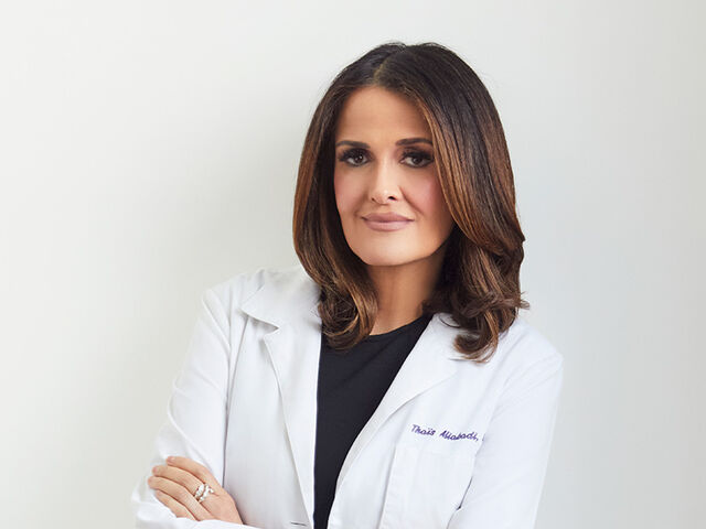 Dr. Thaïs Aliabadi stands against a pale grey studio backdrop in a white lab coat over a black dress, arms folded, looking at the camera.