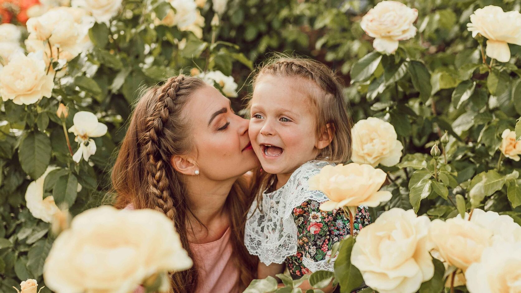 A mother with braided hair kisses her laughing young daughter on the cheek, the two of them surrounded by pale yellow garden roses.