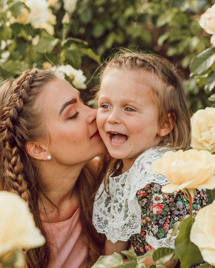 A mother with braided hair kisses her laughing young daughter on the cheek, the two of them surrounded by pale yellow garden roses.