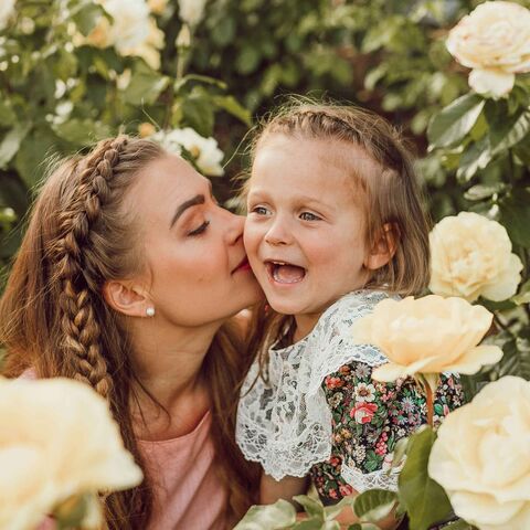 A mother with braided hair kisses her laughing young daughter on the cheek, the two of them surrounded by pale yellow garden roses.
