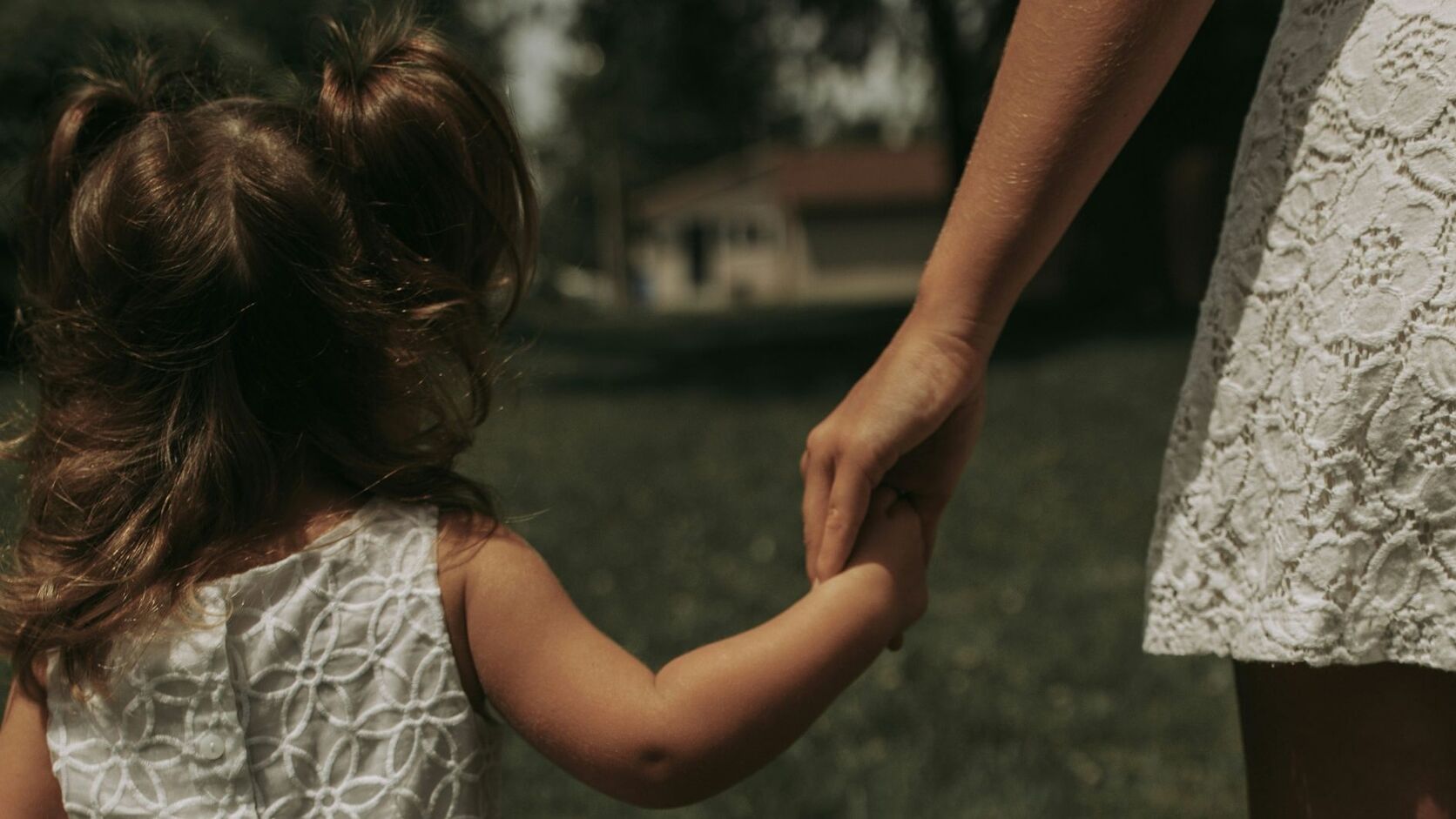 A young girl in a white eyelet dress holds her mother's hand as they walk together across a sunlit lawn, seen from behind.