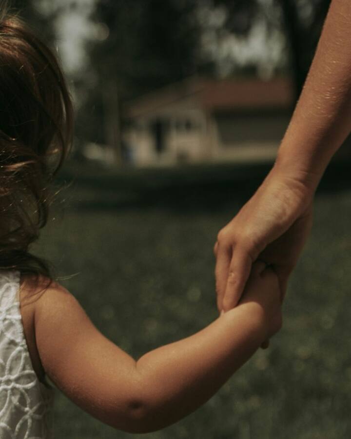 A young girl in a white eyelet dress holds her mother's hand as they walk together across a sunlit lawn, seen from behind.