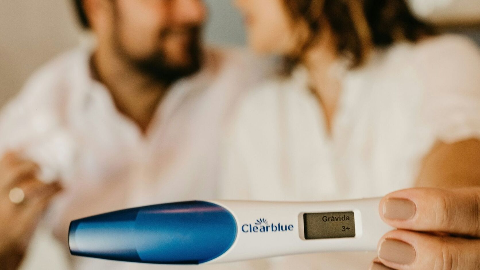 A couple in white sit close together, slightly out of focus, holding a positive digital pregnancy test toward the camera.