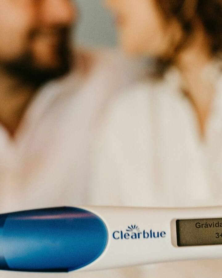A couple in white sit close together holding a positive digital pregnancy test toward the camera.