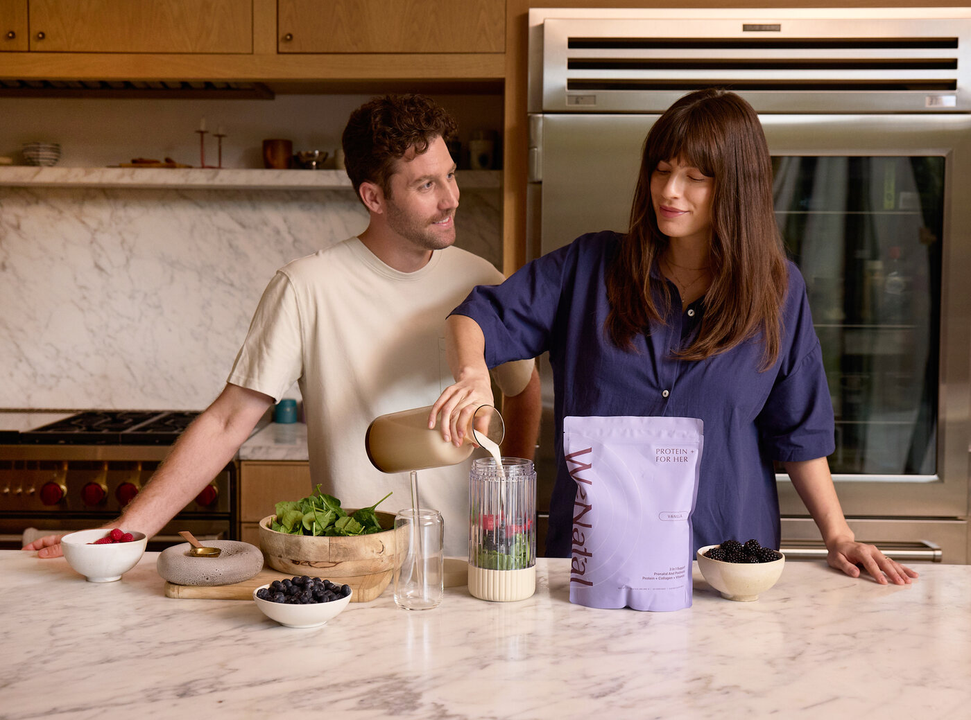 A couple in a marble kitchen makes a smoothie together, pouring WeNatal protein into a blender beside bowls of berries and spinach.