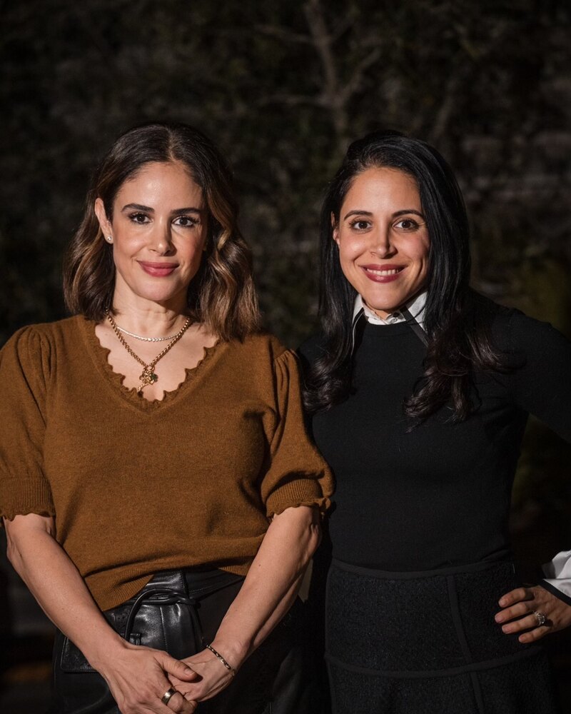 WeNatal co-founders Ronit Menashe and Vida Delrahim stand together, smiling, against a dark garden backdrop.
