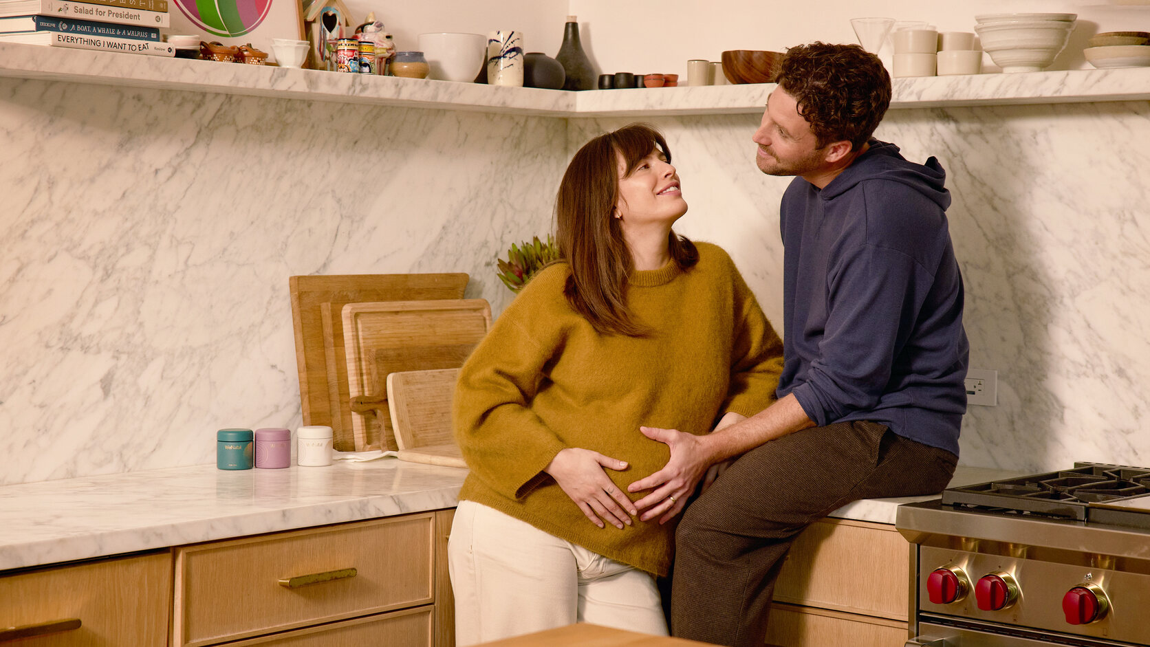A pregnant woman in a mustard sweater and her partner share a tender moment in their marble kitchen, his hand resting on her belly, with WeNatal jars on the counter.