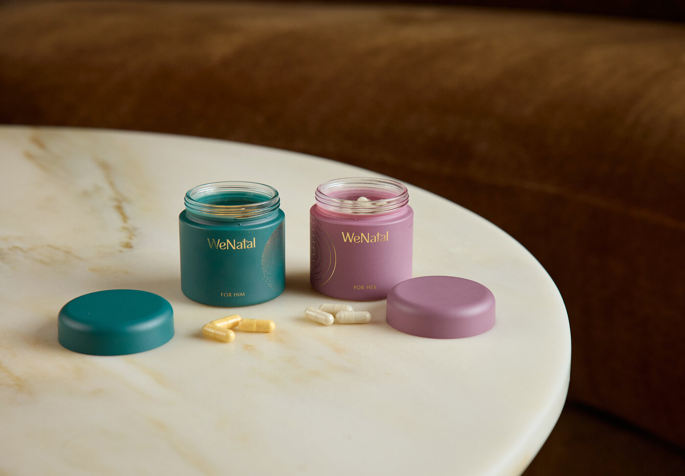 Two open frosted WeNatal jars — a teal 'For Him' and a pink 'For Her' — on a marble table with capsules spilled beside them.