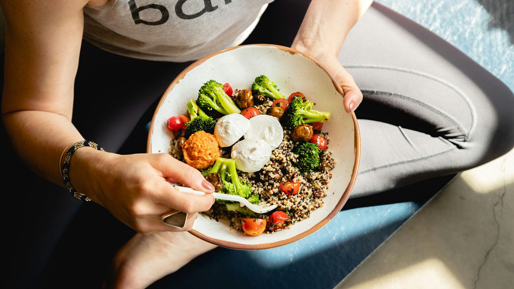 A woman sitting cross-legged holds a grain bowl of quinoa, broccoli, tomatoes, and mozzarella in her lap.