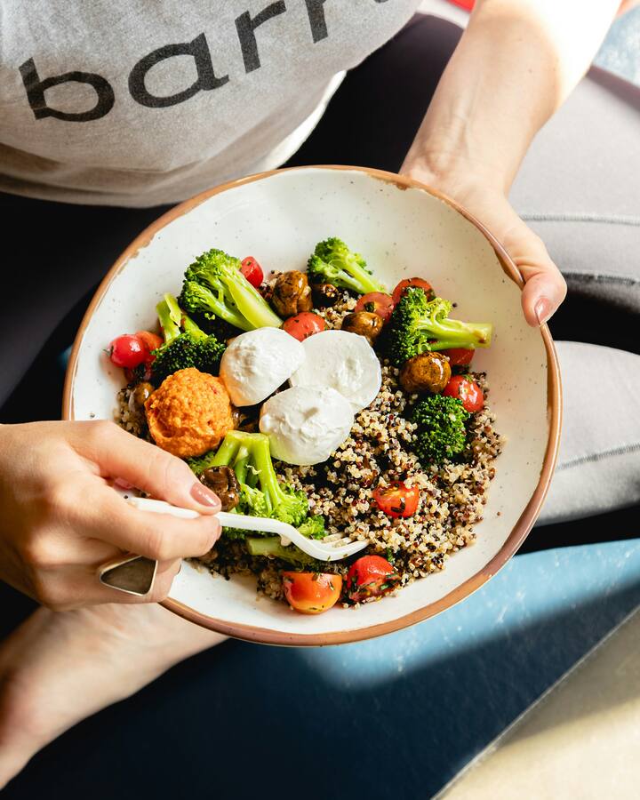 A woman sitting cross-legged holds a grain bowl of quinoa, broccoli, tomatoes, and mozzarella in her lap.