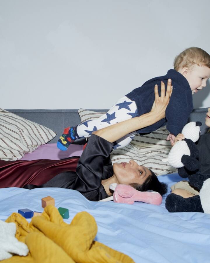 A mother lies back on a bed lifting her baby into the air while her young daughter sits beside her holding a stuffed toy, the bed scattered with soft toys.