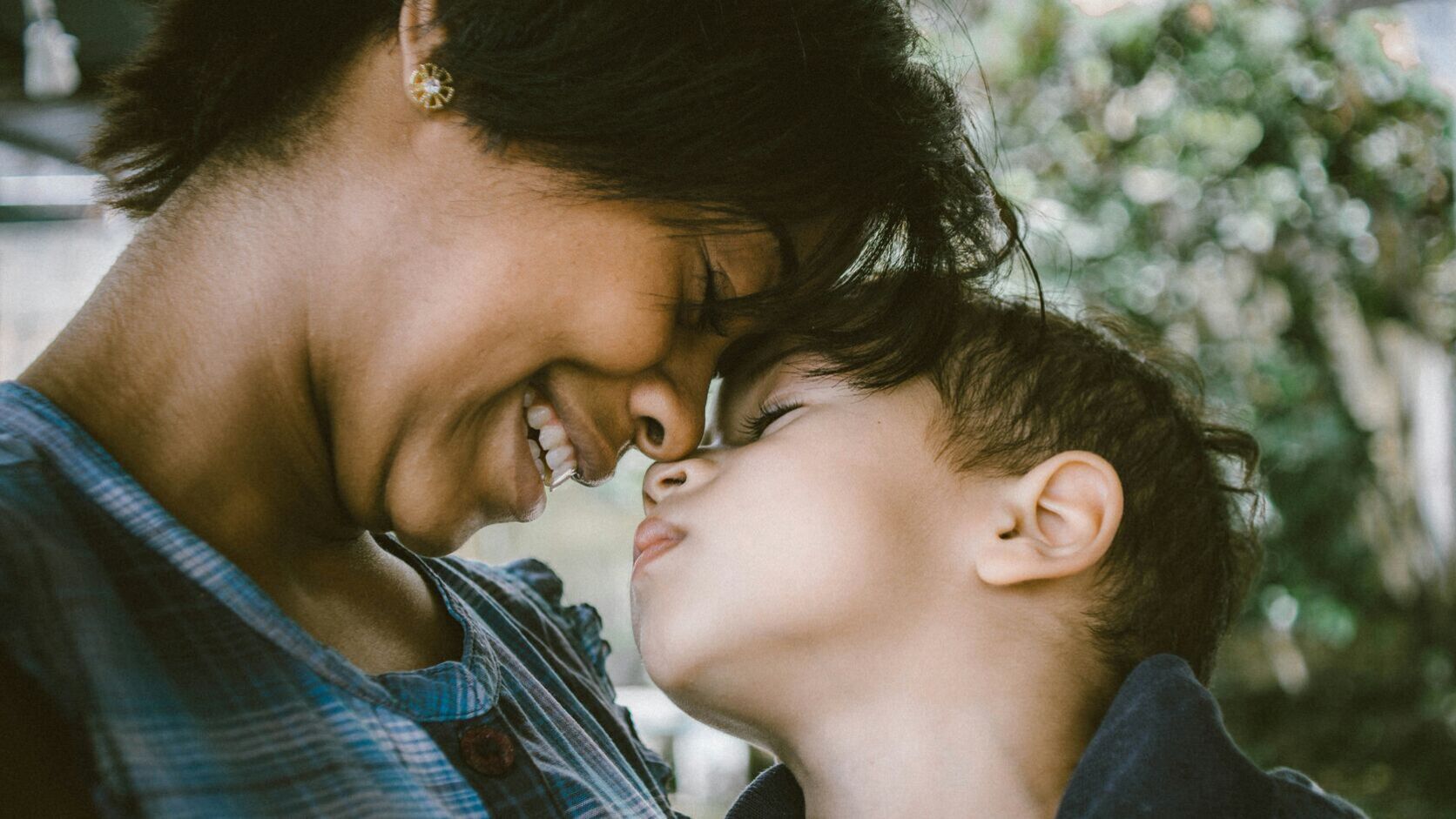A mother in a blue plaid shirt closes her eyes and smiles as she presses her forehead and nose to her young son&rsquo;s, the two of them cheek to cheek outdoors in soft green light.