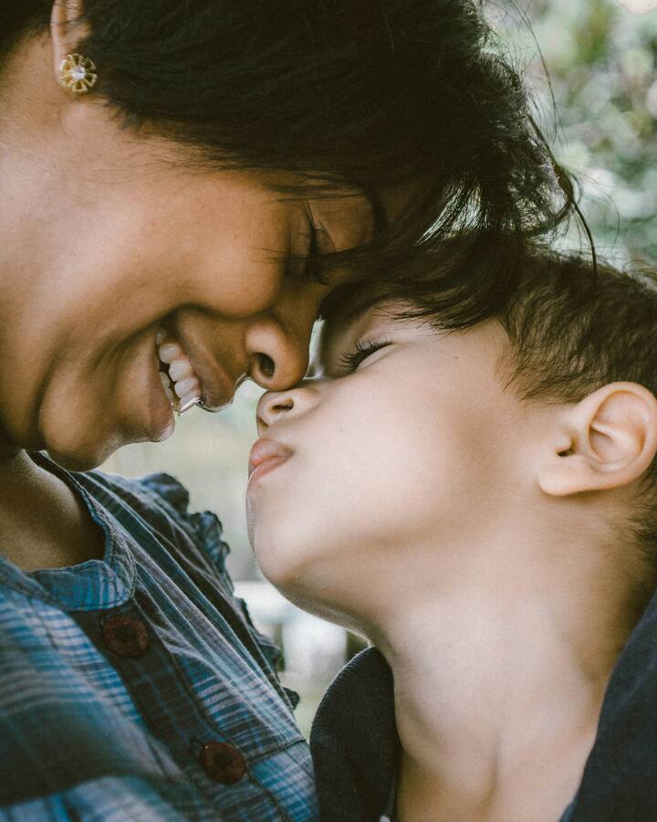 A mother in a blue plaid shirt closes her eyes and smiles as she presses her forehead and nose to her young son&rsquo;s, the two of them cheek to cheek outdoors in soft green light.