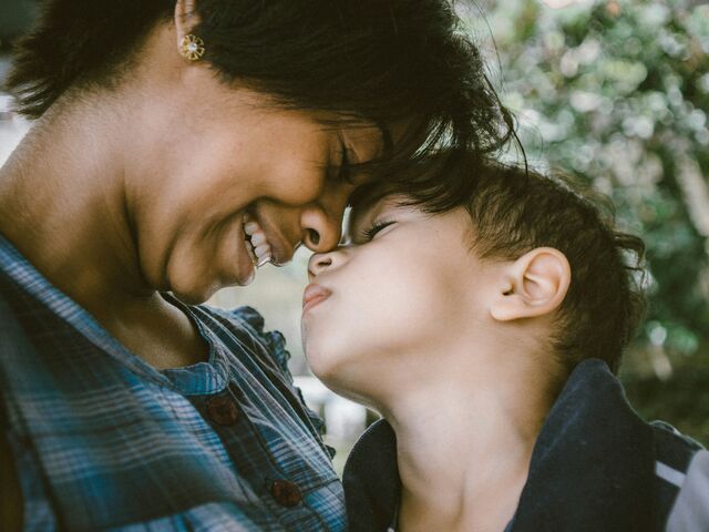 A mother in a blue plaid shirt closes her eyes and smiles as she presses her forehead and nose to her young son&rsquo;s, the two of them cheek to cheek outdoors in soft green light.