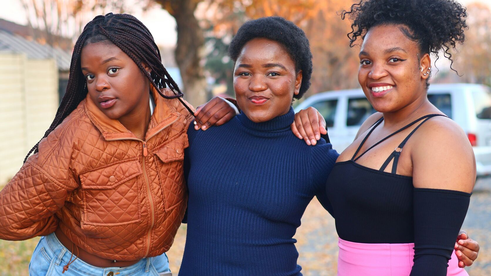 Three teenage girls stand arm in arm on a tree-lined street in autumn, smiling at the camera.