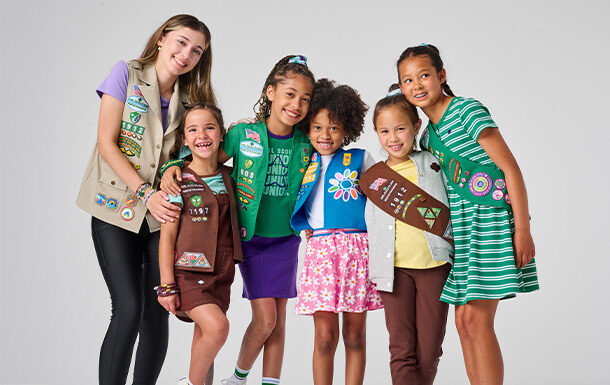 Six Girl Scouts of different ages stand together in their Daisy, Brownie, Junior, Cadette, Senior, and Ambassador uniforms against a pale grey backdrop.