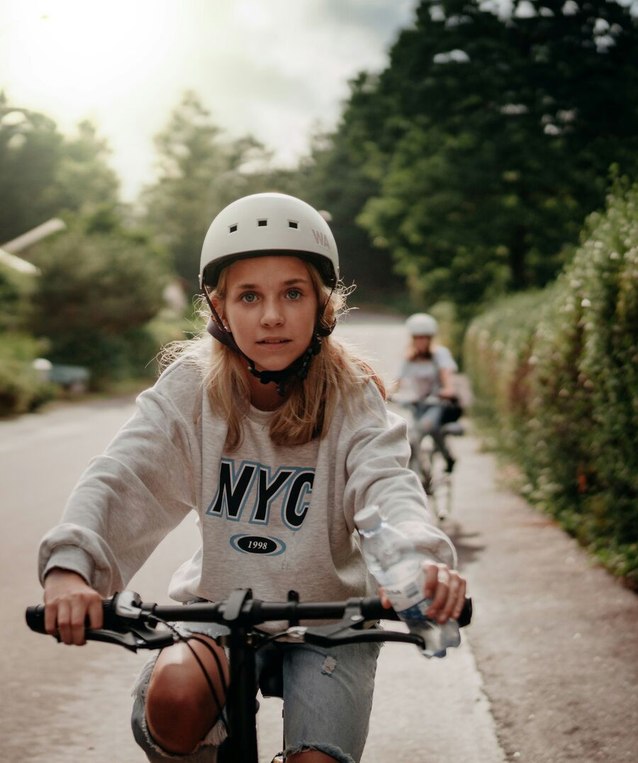 A girl in a white helmet and grey NYC sweatshirt rides her bike down a hedge-lined lane, a friend following behind her.