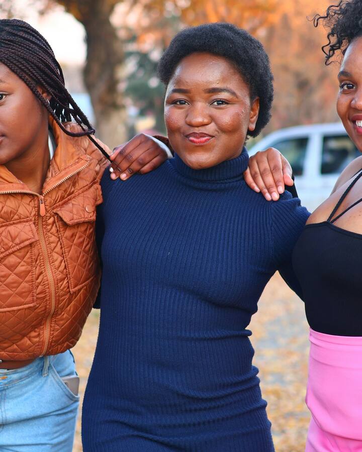 Three teenage girls stand arm in arm on a tree-lined street in autumn, smiling at the camera.