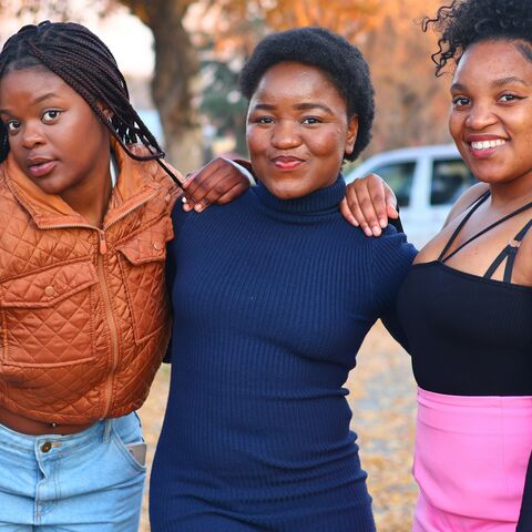 Three teenage girls stand arm in arm on a tree-lined street in autumn, smiling at the camera.