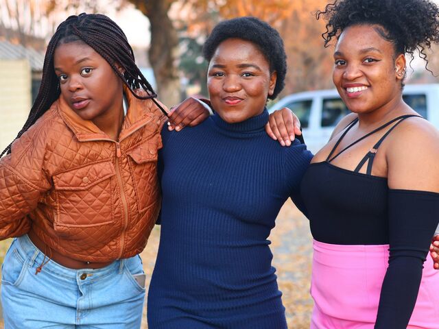 Three teenage girls stand arm in arm on a tree-lined street in autumn, smiling at the camera.