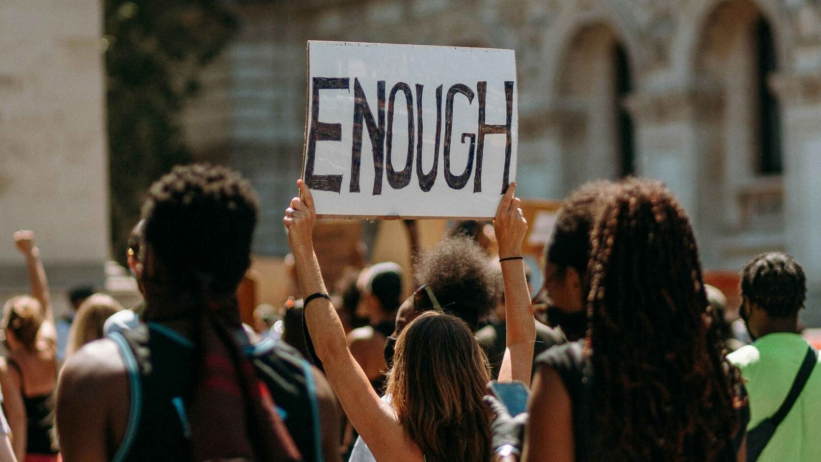 A protester at a crowded outdoor rally holds a hand-lettered cardboard sign reading 'ENOUGH' above the crowd.