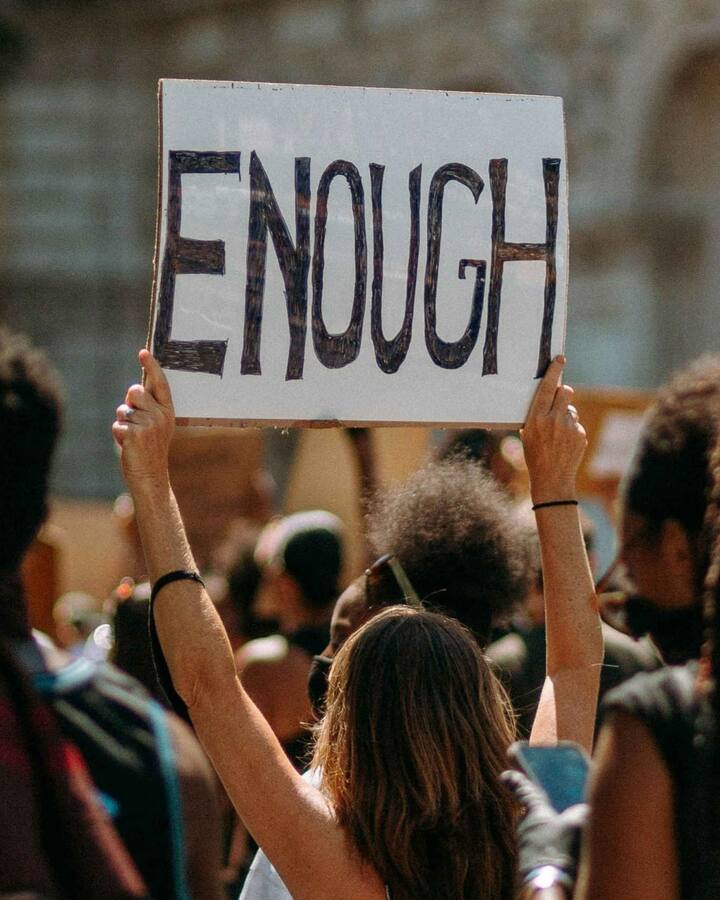 A protester at a crowded outdoor rally holds a hand-lettered cardboard sign reading 'ENOUGH' above the crowd.