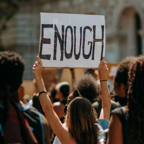 A protester at a crowded outdoor rally holds a hand-lettered cardboard sign reading 'ENOUGH' above the crowd.