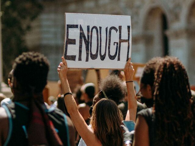 A protester at a crowded outdoor rally holds a hand-lettered cardboard sign reading 'ENOUGH' above the crowd.