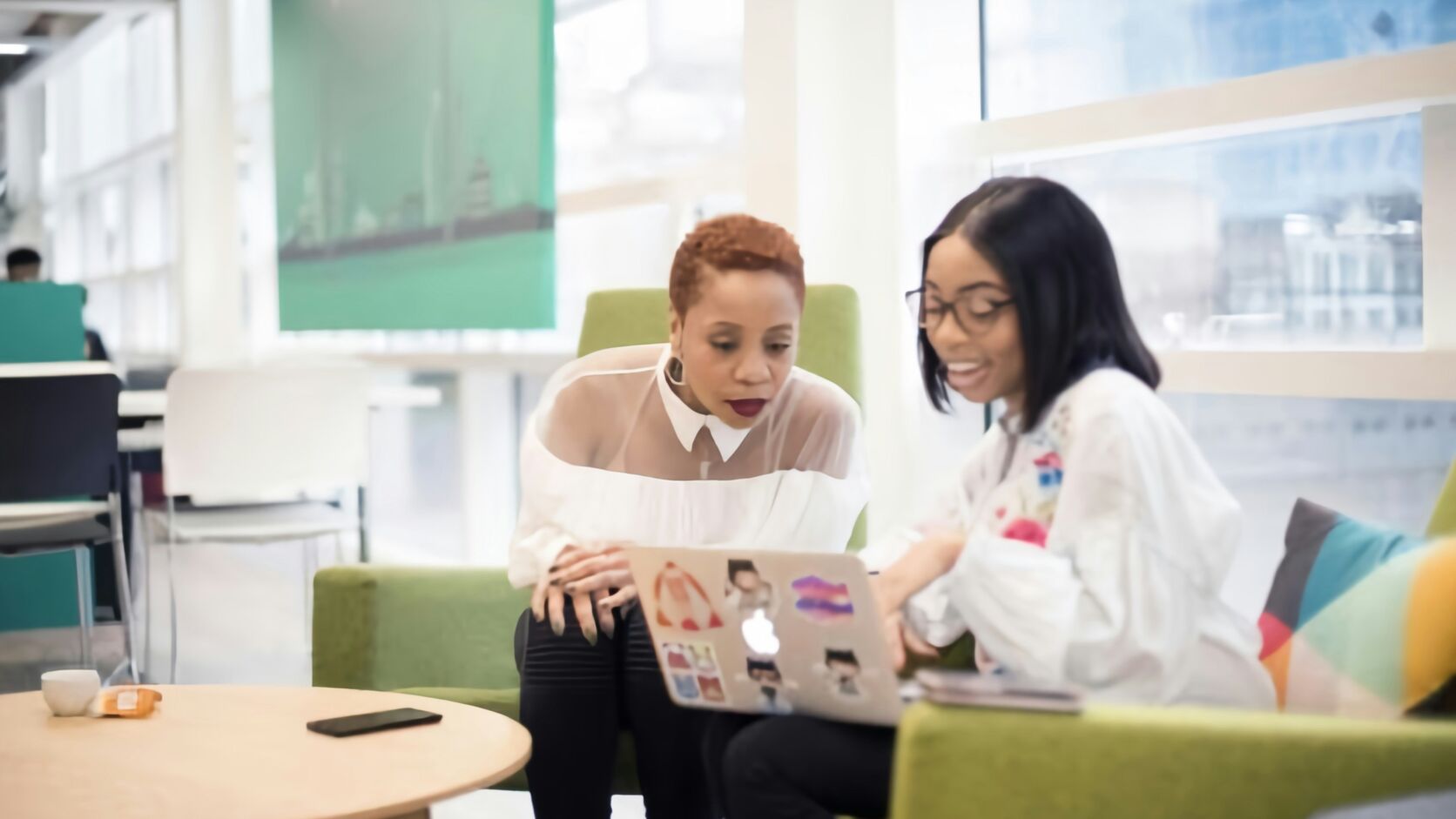 Two Black women professionals sit together on green sofas in a bright modern office, looking at a laptop mid-conversation.