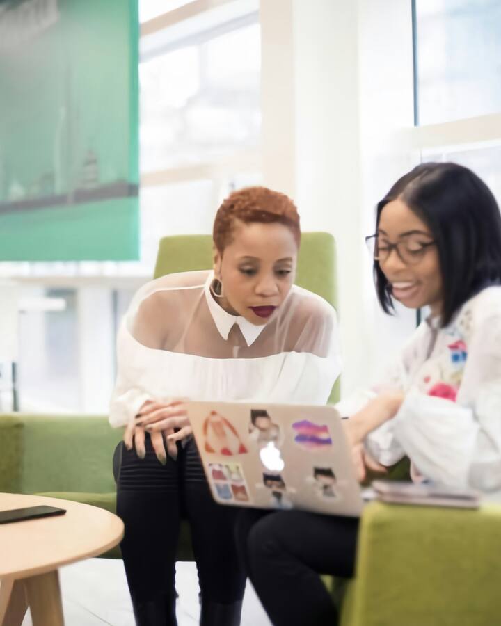 Two Black women professionals sit together on green sofas in a bright modern office, looking at a laptop mid-conversation.